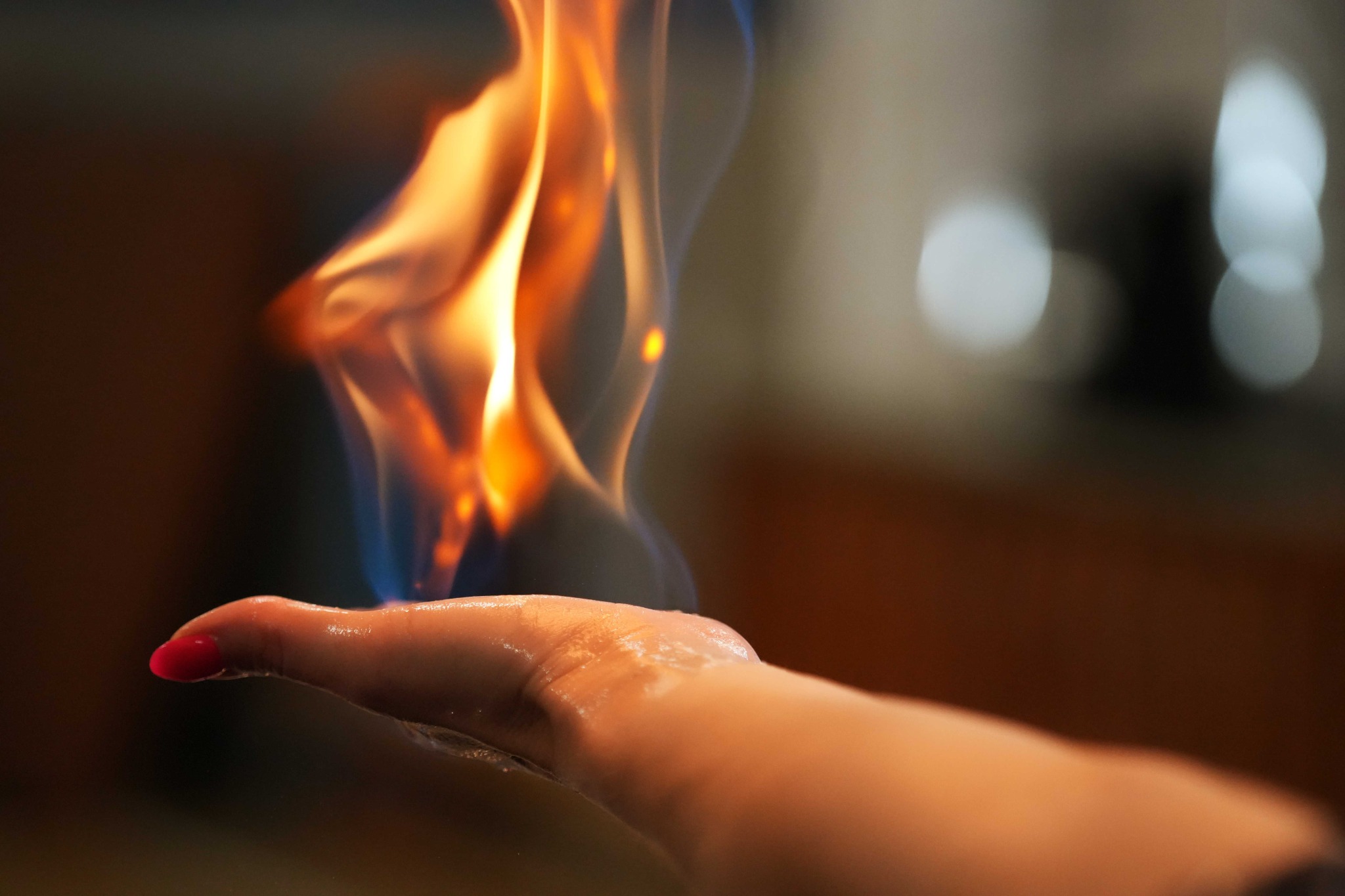 Płomień delikatnie unoszący się nad otwartą dłonią z czerwonym lakierem na paznokciu, na rozmytym tle.