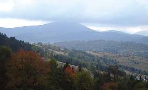 Pejzaż górski w pochmurny dzień. W dolinie widoczne drzewa, niektóre z jesiennymi, pomarańczowymi liśćmi. W tle zalesione zbocza i szczyt góry spowity mgłą.