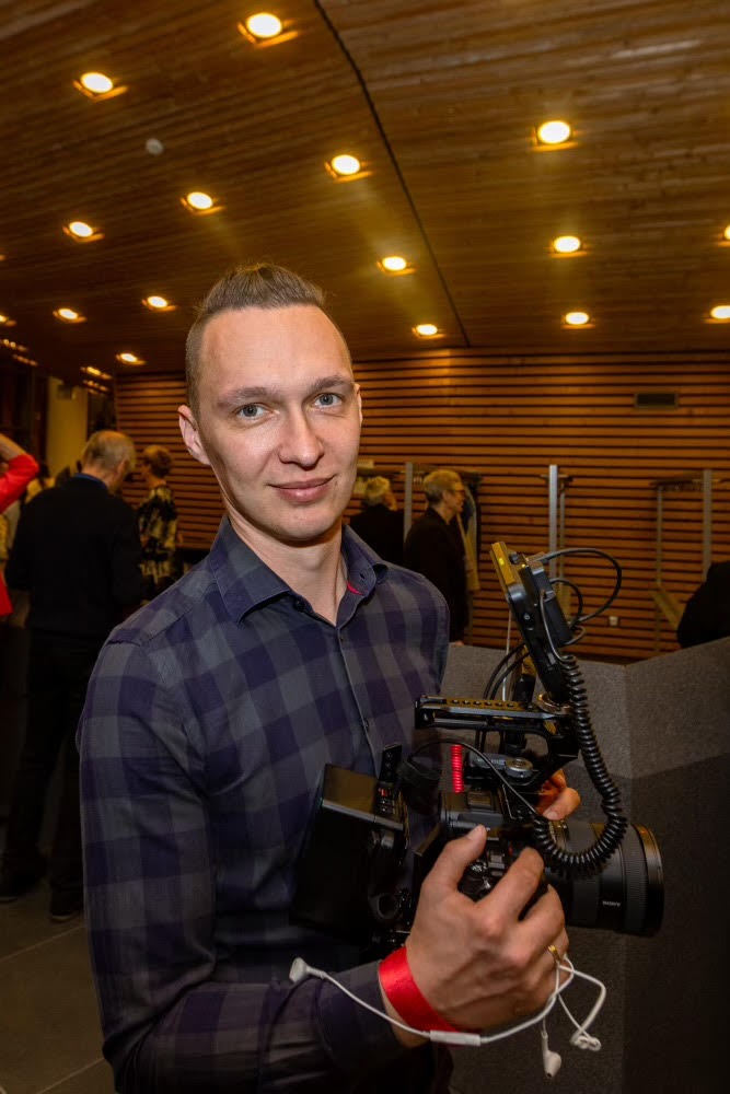 Uśmiechnięty kamerzysta z profesjonalnym sprzętem, w tle drewniana ściana i sufit z oświetleniem punktowym. Widoczny czerwony pasek na nadgarstku.