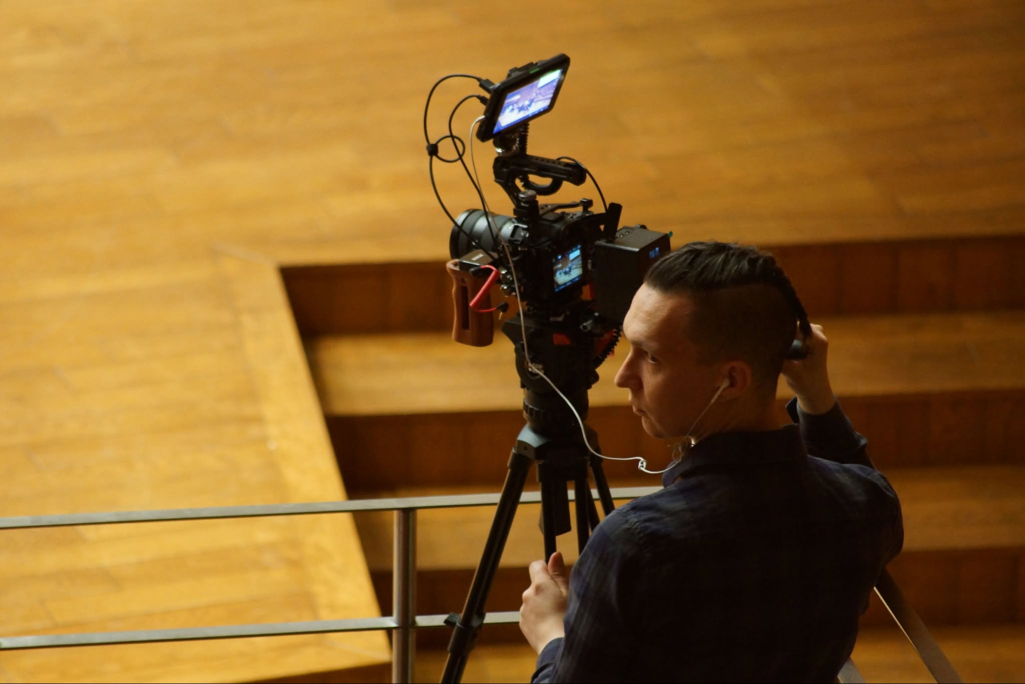 Mężczyzna w słuchawkach ustawia kamerę na statywie przed drewnianą ścianą. Sprzęt z ekranem podglądowym i mikrofonem. Ujęcie z profilu.