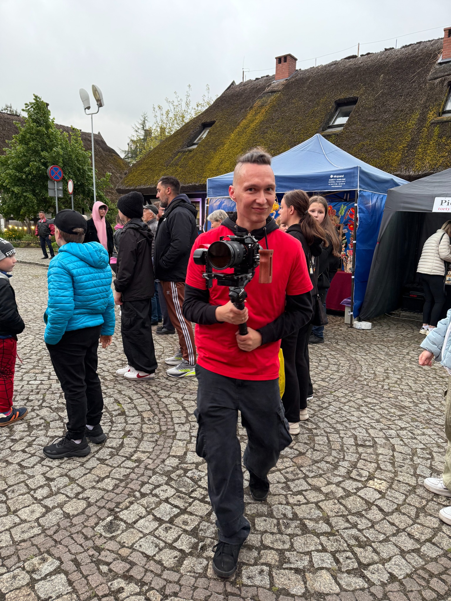 Uśmiechnięty mężczyzna z kamerą na gimbalu, w czerwonej koszulce, na tle straganów i tłumu ludzi na brukowanym placu.