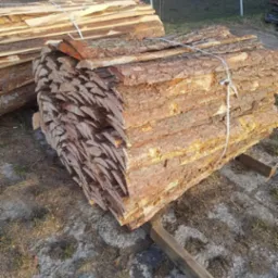 Stosy kory drzewnej związanej sznurkiem, leżące na drewnianych belkach na zewnątrz.