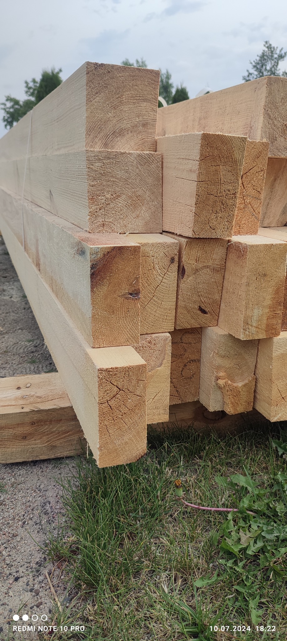 Świeżo przetarte, jasne belki tarcicy budowlanej ułożone w stos na trawie w Kole, widoczne ślady po cięciu.