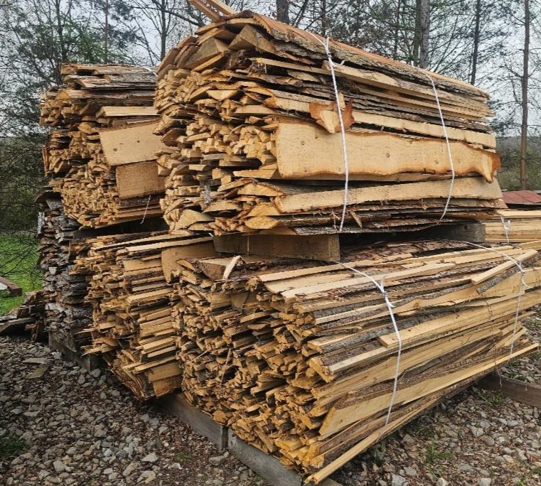 Stosy pociętych desek z korą, spięte sznurkiem, ułożone na paletach na żwirowym podłożu, w tle drzewa.