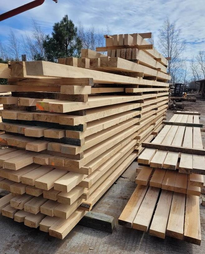 Stosy jasnej, obrobionej tarcicy ułożone na paletach na zewnątrz, widoczny w tle sprzęt do obróbki drewna.
