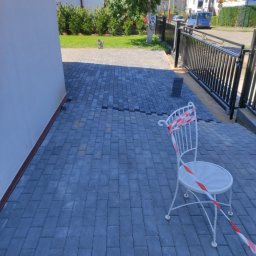 Daminwest - Niedokończone układanie szarej kostki brukowej przed domem, białe krzesło ogrodowe z taśmą ostrzegawczą, widoczne niezakończone fragmenty nawierzchni.