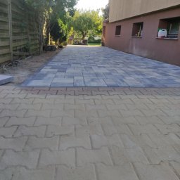 Daminwest - Ułożona kostka brukowa w odcieniach szarości i beżu tworząca ścieżkę wzdłuż budynku, z widocznym fragmentem starej nawierzchni z kostki betonowej.