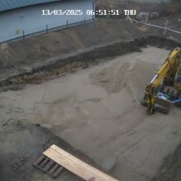 Daminwest - Żółta koparka w wykopie pod fundamenty domu jednorodzinnego. Widok z góry z kamery przemysłowej. W tle budynek i drzewa.