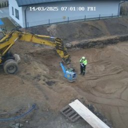 Daminwest - Żółta koparka i pracownik zagęszczają piasek w wykopie pod budowę. W tle budynek i ogrodzenie. Widok z kamery monitoringu z datą 14/03/2025.