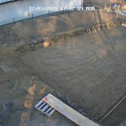 Daminwest - Przygotowany teren pod budowę z wykopem, drewnianym szalunkiem i stosem cegieł, widok z góry z datą i godziną na zdjęciu. Pracownik w odblaskowej kamizelce.