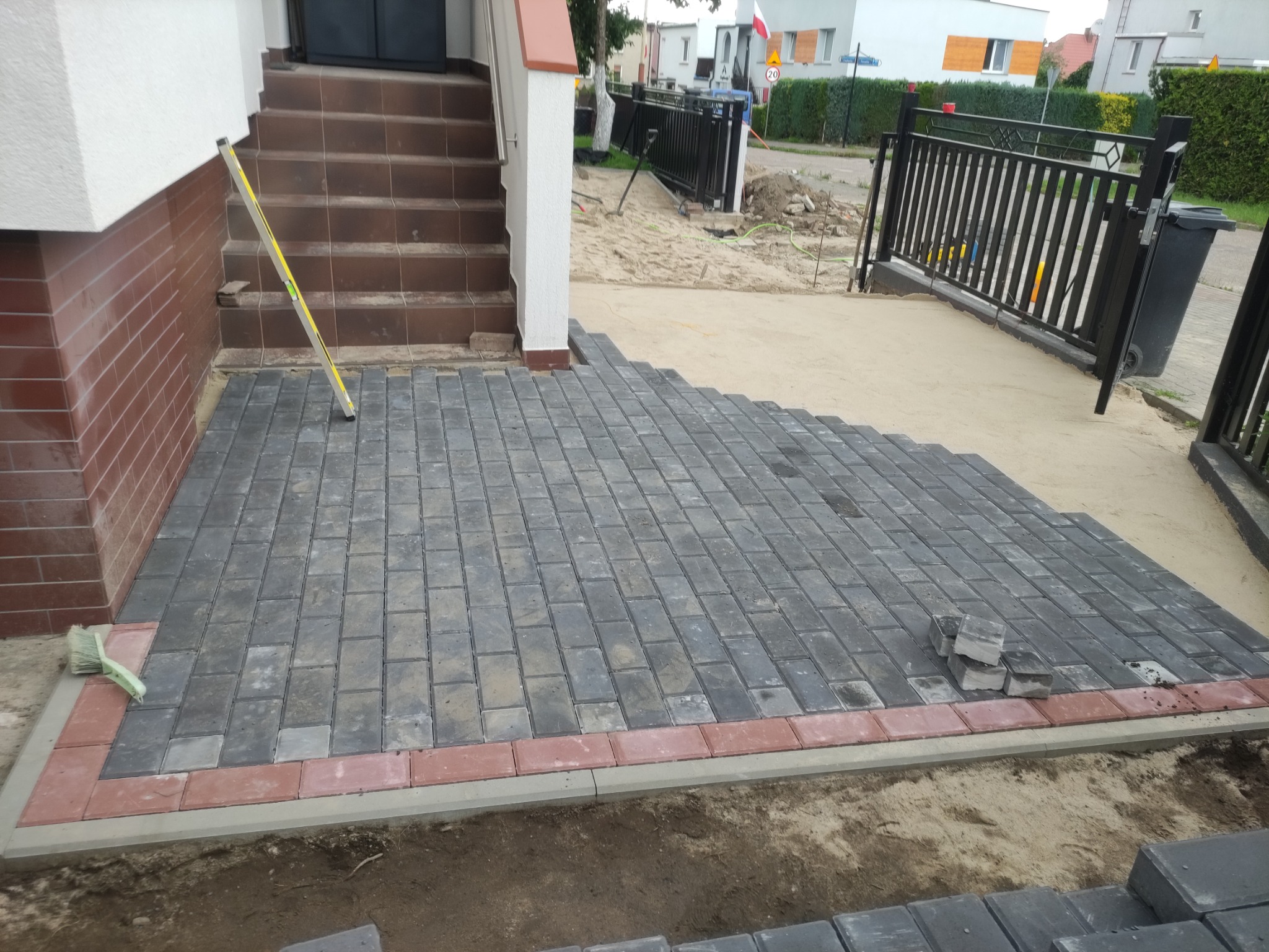 Fragment układanej nawierzchni z szarej kostki brukowej z czerwoną obwódką, widoczny poziomica oparta o ścianę budynku, szczotka do zamiatania i stos kostki w tle.