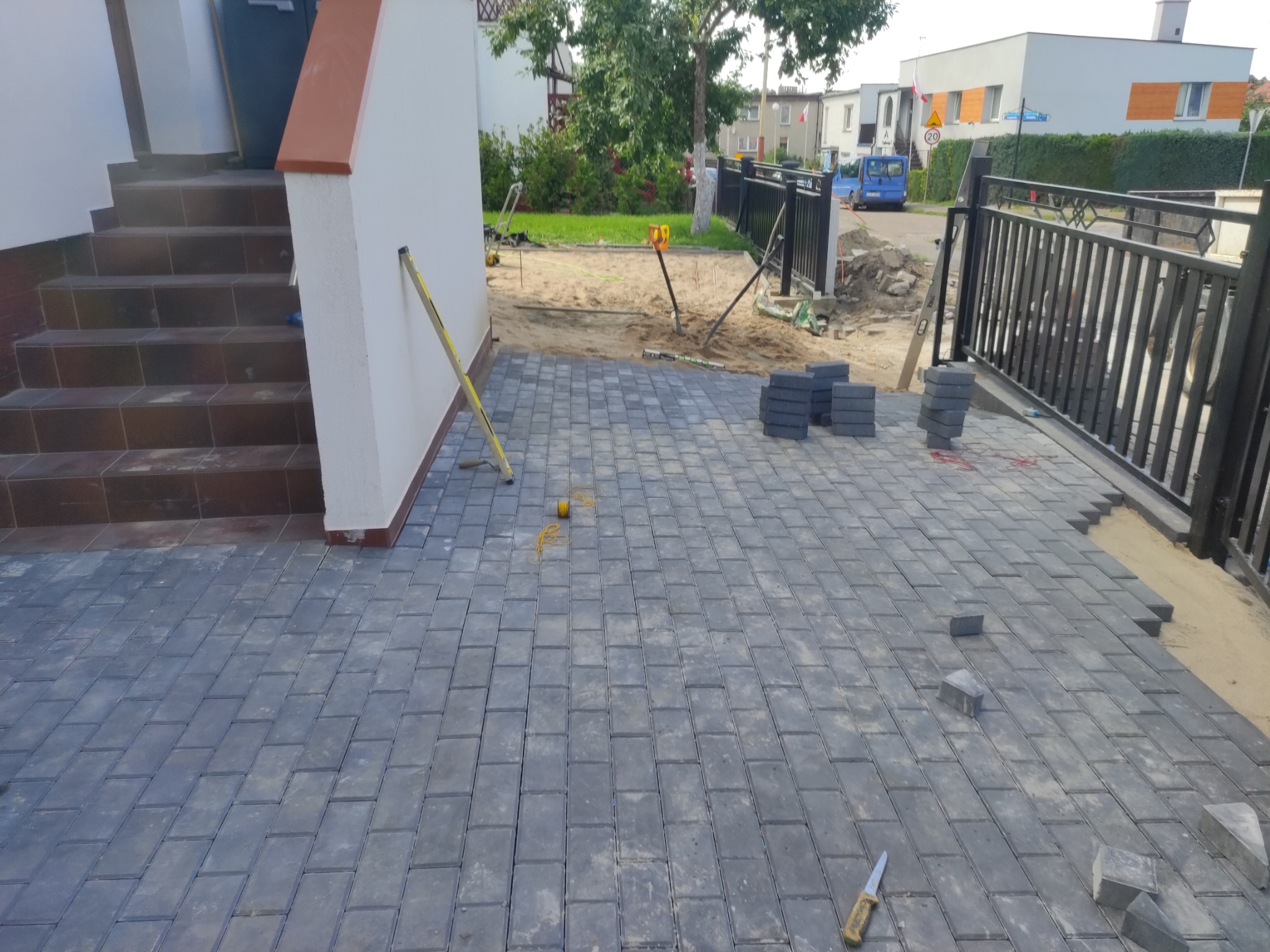 Ułożona częściowo szara kostka brukowa przed domem, widoczny poziomica, nożyk i stosy kostki, teren przygotowany pod dalsze prace.