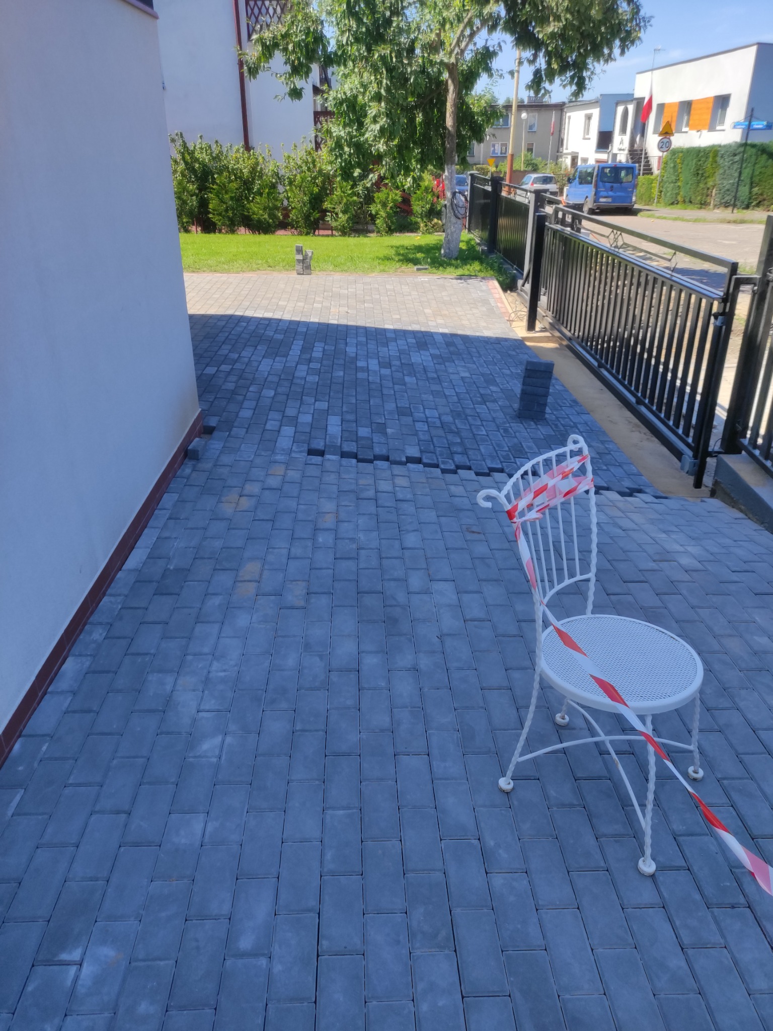 Niedokończone układanie szarej kostki brukowej przed domem, białe krzesło ogrodowe z taśmą ostrzegawczą, widoczne niezakończone fragmenty nawierzchni.