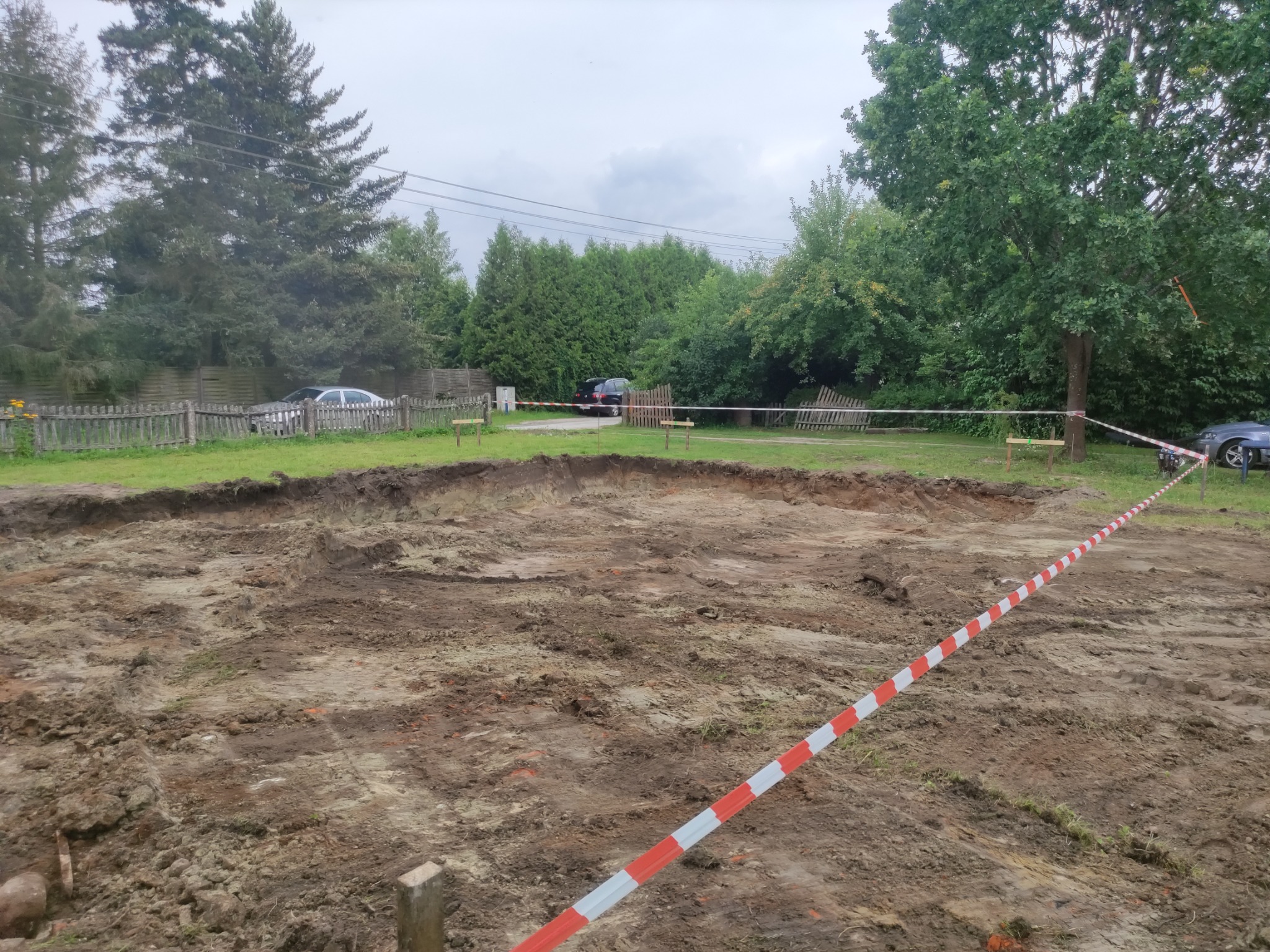 Wykonany wykop w ziemi otoczony taśmą ostrzegawczą, drzewa w tle i fragmenty ogrodzenia.
