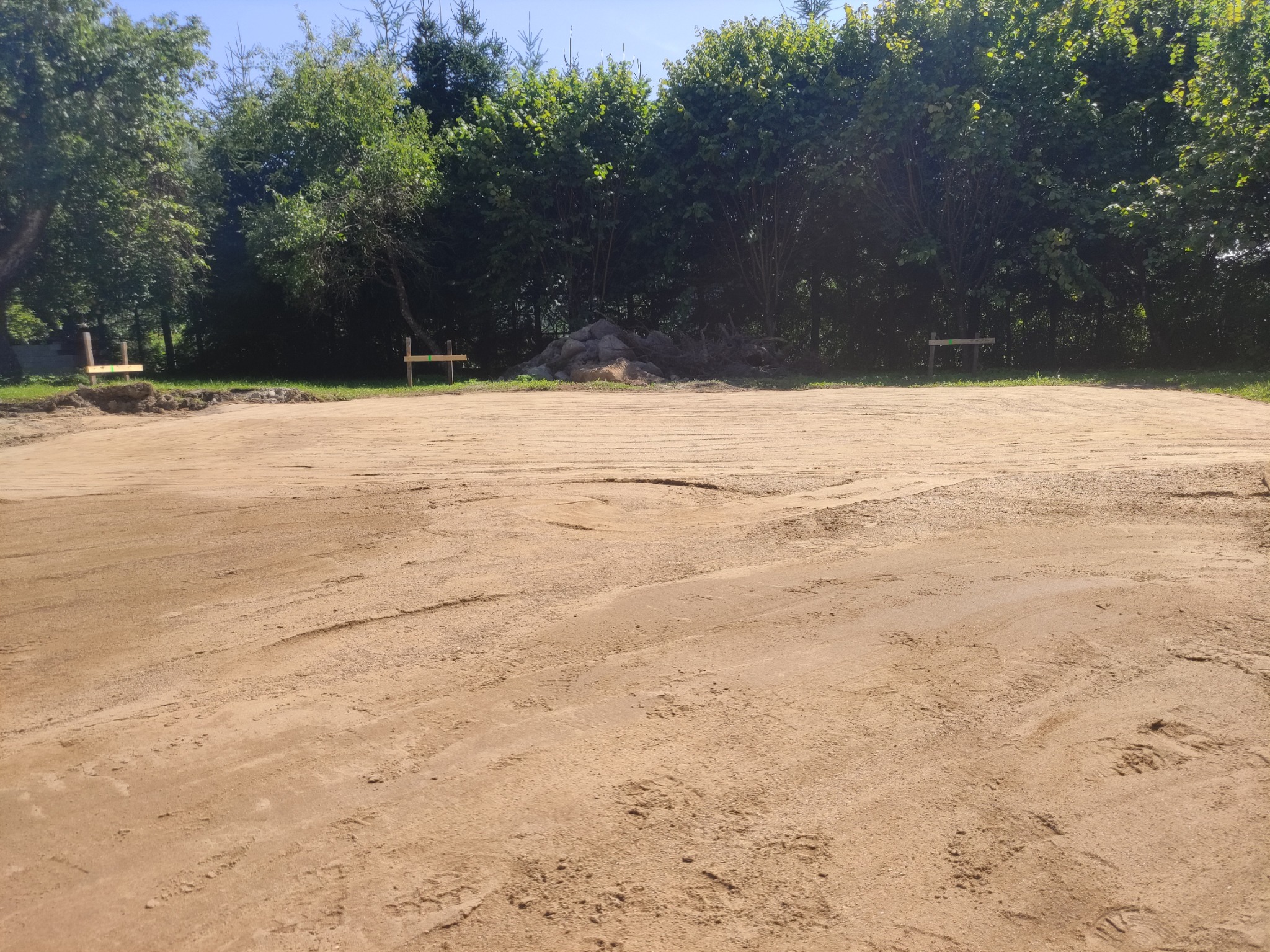 Przygotowany teren pod budowę, wyrównana piaszczysta powierzchnia z widocznymi śladami opon, w tle drzewa i pryzma ziemi.