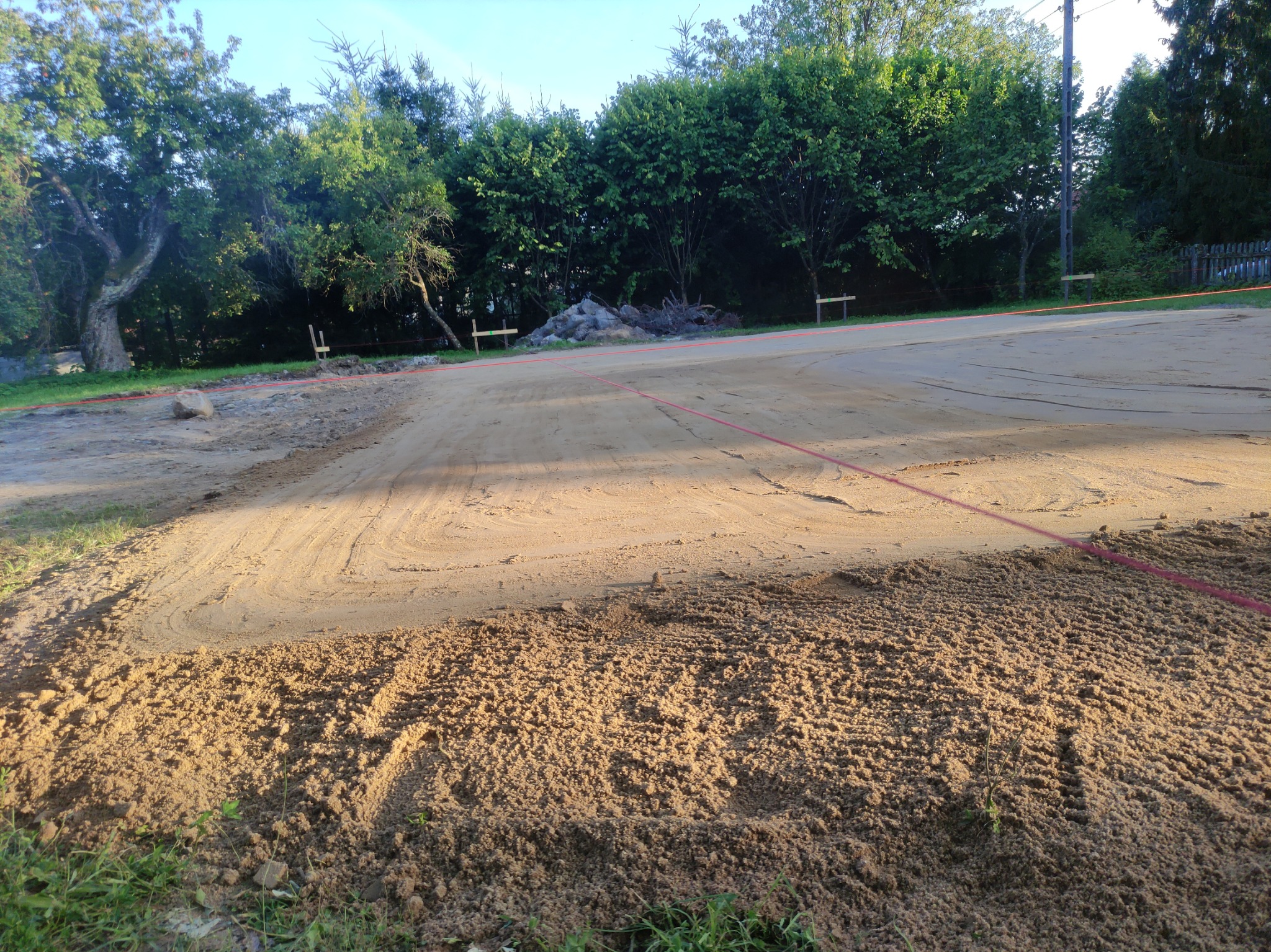 Przygotowany teren pod budowę, wyrównany piasek z widocznymi śladami opon, wytyczony sznurkiem, w tle zieleń drzew i krzewów.