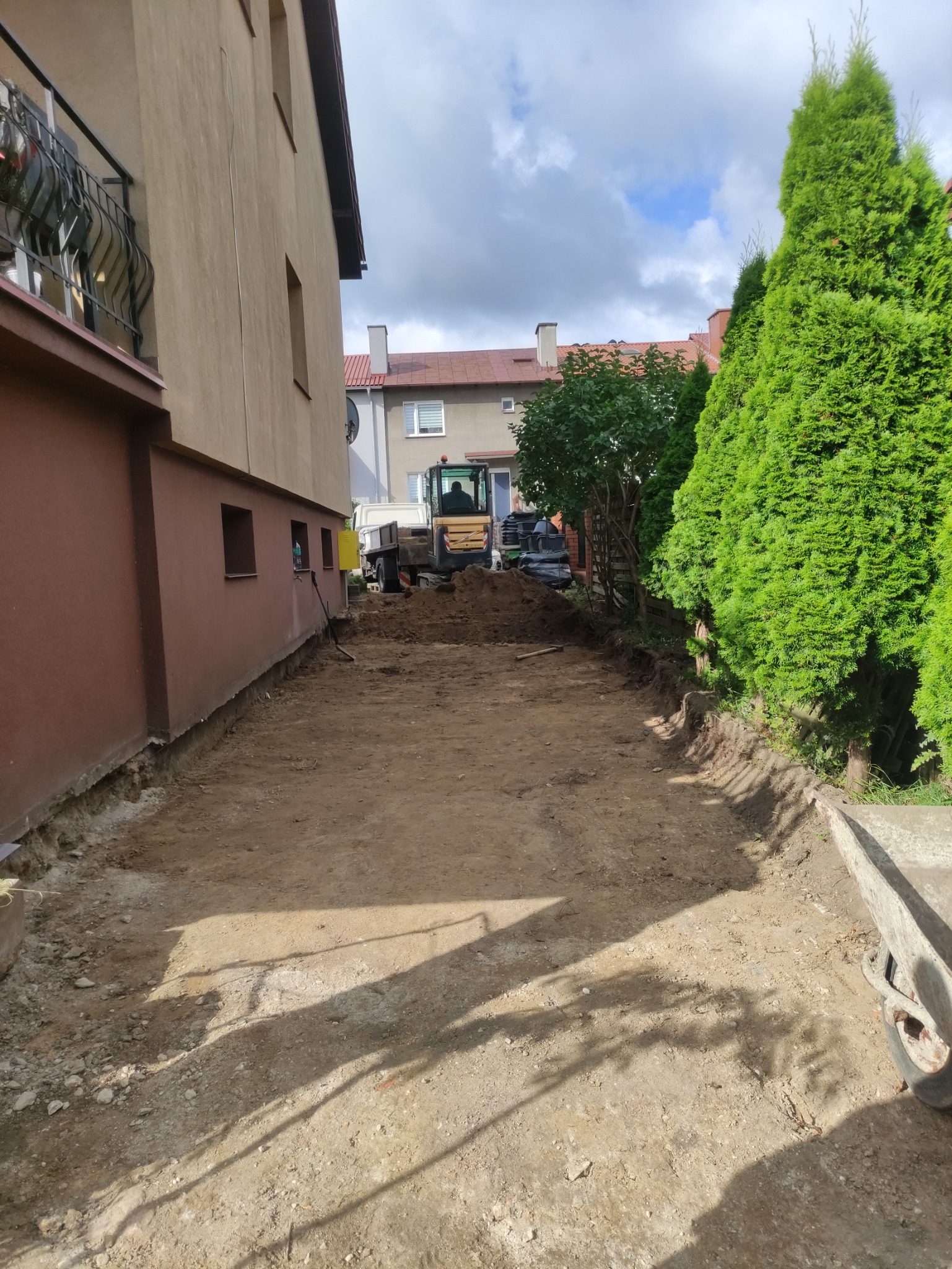 Przygotowany teren pod budowę z widocznym wykopem, piaskiem i maszyną budowlaną w tle, sąsiedztwo budynków mieszkalnych i iglaków.