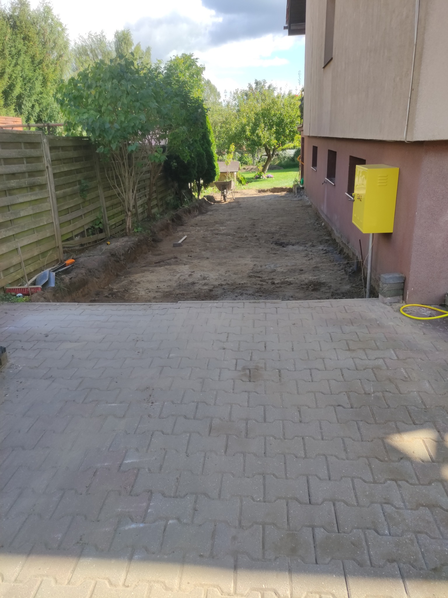 Przygotowany teren pod budowę lub układanie kostki brukowej, widoczny wykop wzdłuż ogrodzenia i budynku, częściowo ułożona kostka brukowa w pierwszym planie, żółta skrzynka gazowa na ścianie budynku.