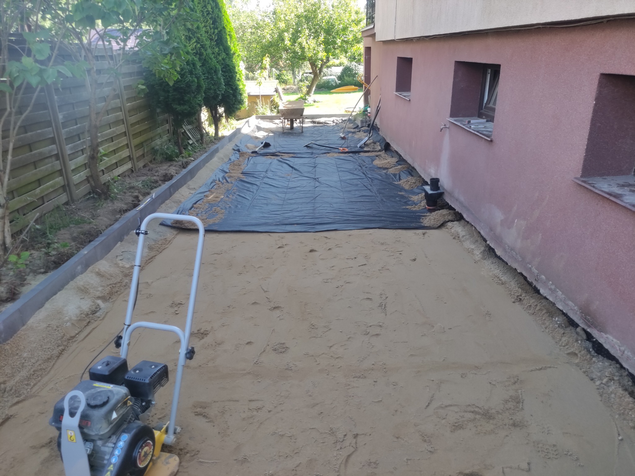 Przygotowanie terenu pod kostkę brukową: ubijarka wibracyjna na piasku, obrzeża ułożone wzdłuż ogrodzenia i ściany budynku, czarna geowłóknina częściowo rozłożona, taczka i narzędzia w tle.