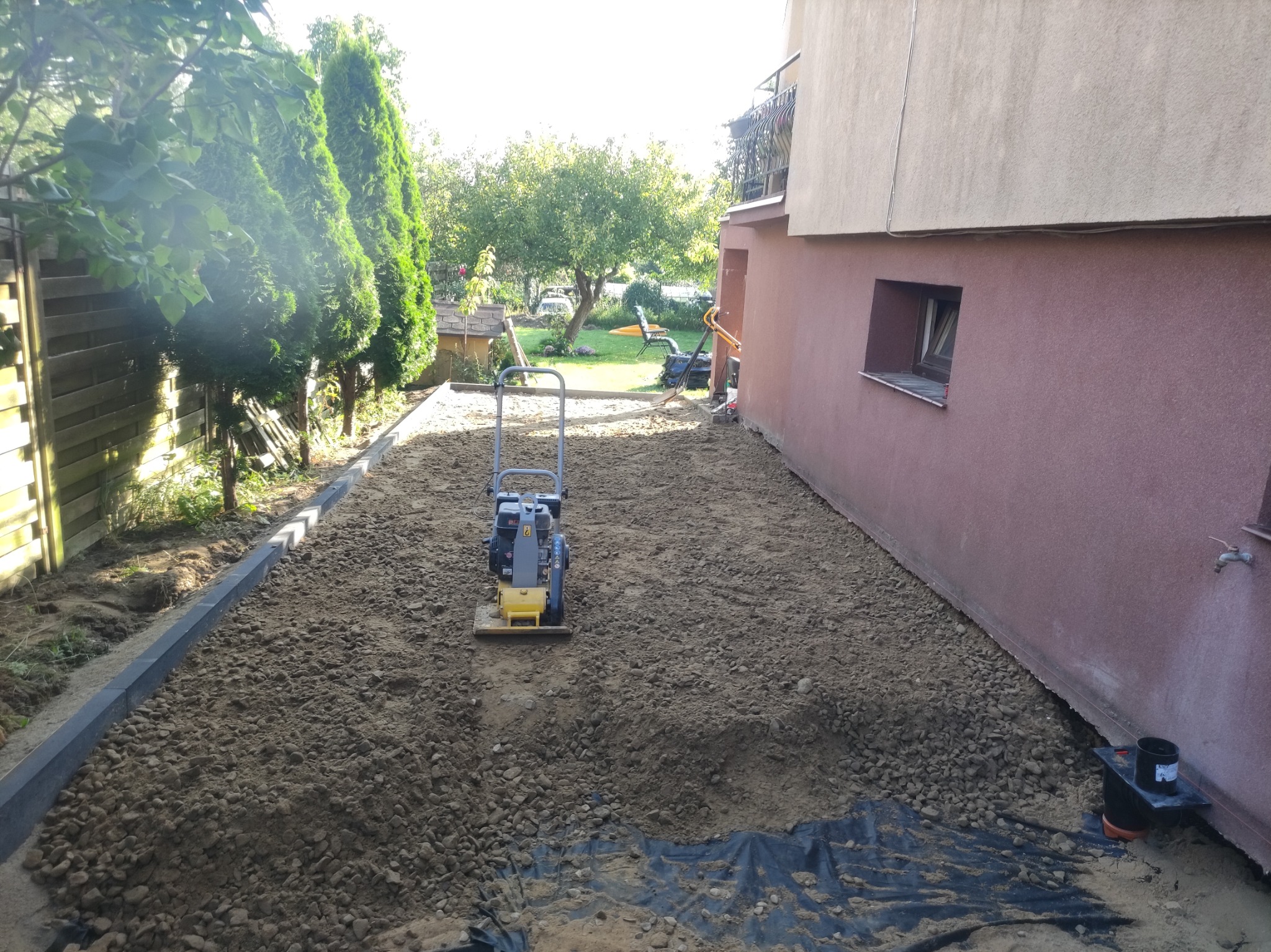 Przygotowanie podłoża pod kostkę brukową z użyciem zagęszczarki w ogrodzie przydomowym, widoczne obrzeża i warstwa podsypki.