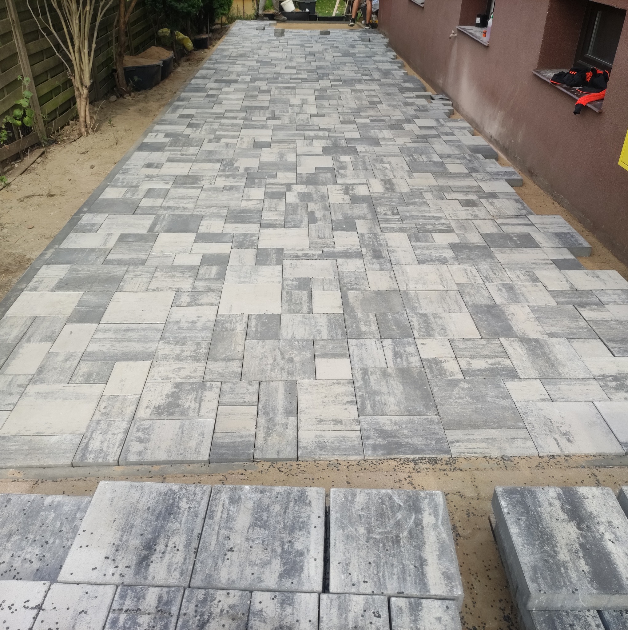 Ułożona ścieżka z szaro-białej kostki brukowej o nieregularnym wzorze, z widocznymi zapasami kostki i fragmentem budynku w tle.