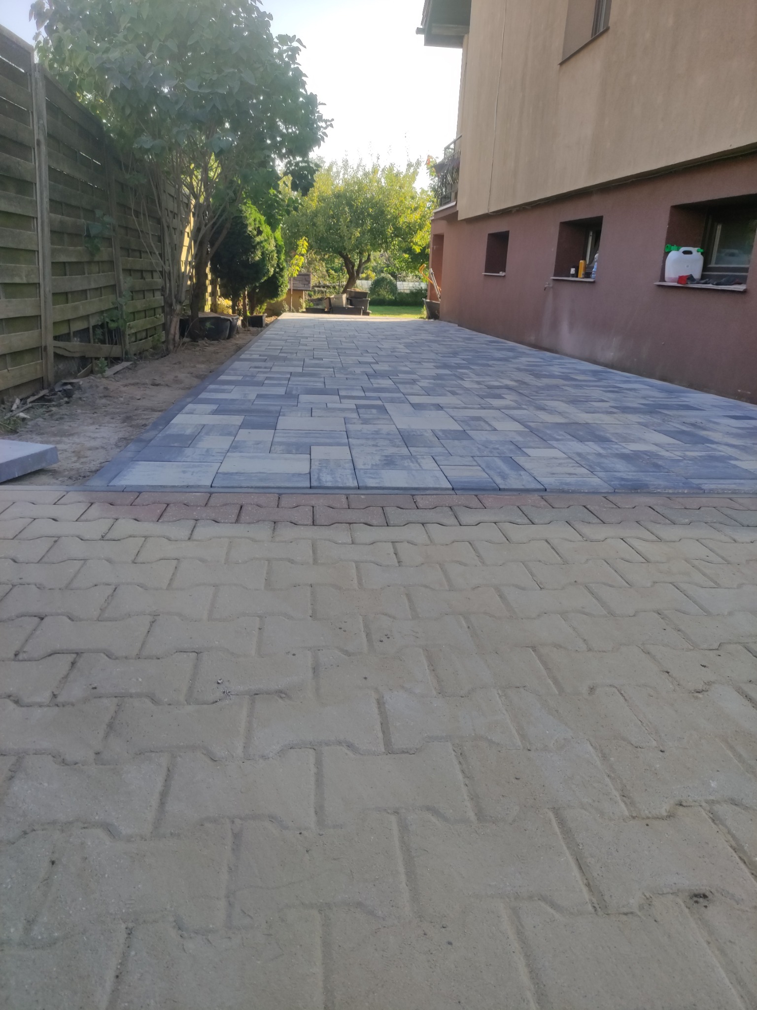 Ułożona kostka brukowa w odcieniach szarości i beżu tworząca ścieżkę wzdłuż budynku, z widocznym fragmentem starej nawierzchni z kostki betonowej.