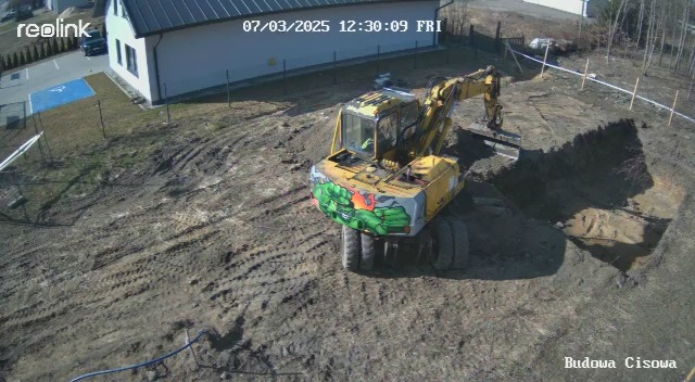 Żółta koparka z grafiką na tle wykopu i budynku z rampą dla niepełnosprawnych. Zdjęcie z kamery przemysłowej.