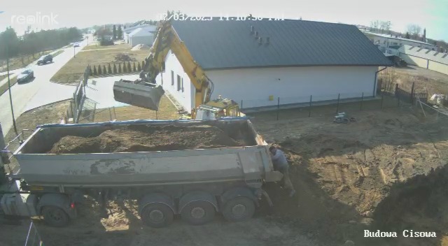 Żółta koparka ładuje ziemię do ciężarówki na tle budynku. Widoczny pracownik przy ciężarówce i otoczenie placu budowy. Znak wodny 'Budowa Cisowa'.