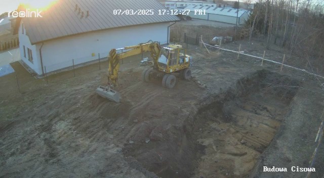 Żółta koparka kołowa w trakcie prac ziemnych na budowie, widok z góry. Wykop pod fundamenty, ogrodzenie z taśmy wygradzającej. Dom w tle, wczesny etap budowy.