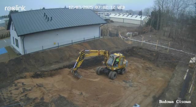 Żółta koparka w trakcie prac ziemnych na budowie, widok z góry, data i logo kamery w rogu.