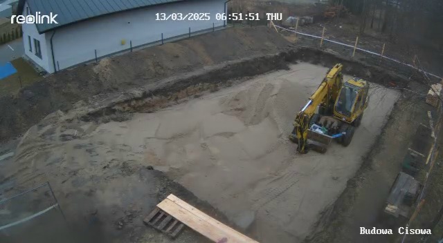 Żółta koparka w wykopie pod fundamenty domu jednorodzinnego. Widok z góry z kamery przemysłowej. W tle budynek i drzewa.