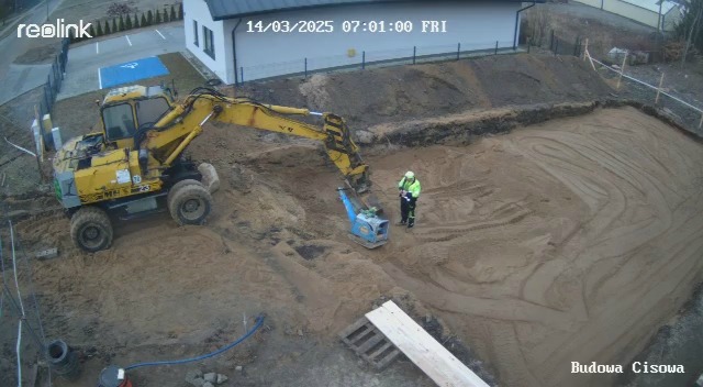 Żółta koparka i pracownik zagęszczają piasek w wykopie pod budowę. W tle budynek i ogrodzenie. Widok z kamery monitoringu z datą 14/03/2025.