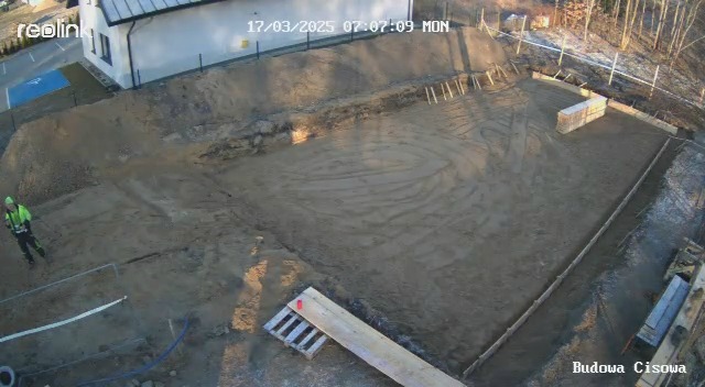 Przygotowany teren pod budowę z wykopem, drewnianym szalunkiem i stosem cegieł, widok z góry z datą i godziną na zdjęciu. Pracownik w odblaskowej kamizelce.