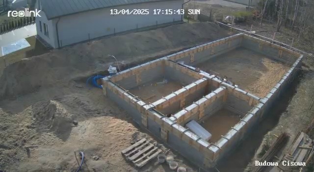 Zalane fundamenty z drewnianym szalunkiem, widok z góry. Na ziemi leżą narzędzia i materiały budowlane. W tle widać budynek i drzewa.