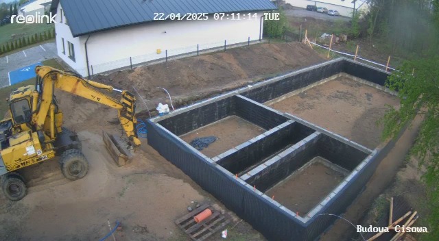 Żółta koparka przy wykopie pod fundamenty z izolacją, widok z góry. Na drugim planie budynek z szarym dachem i trawnik.