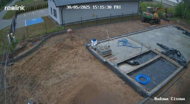 Budowa w toku: robotnik zagęszcza piasek obok fundamentów, widoczny wykop i maszyna budowlana w tle, nagranie z kamery przemysłowej z datą.