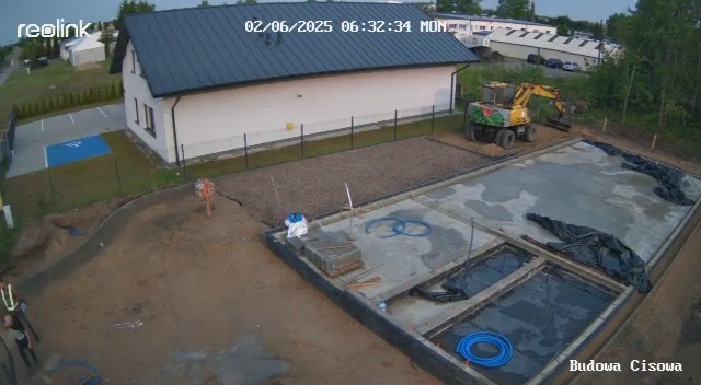 Budowa domu z żółtą koparką w tle. Widoczne fundamenty, teren przygotowany pod kostkę. Ujęcie z góry, w tle budynek z szarym dachem i parking.