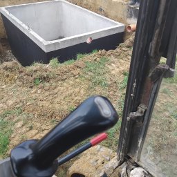 SILWOJ - Betonowy zbiornik w wykopie, widok z kabiny koparki. Zbiornik zabezpieczony czarną powłoką, rura w ścianie. W tle ziemia i fragmenty osób.