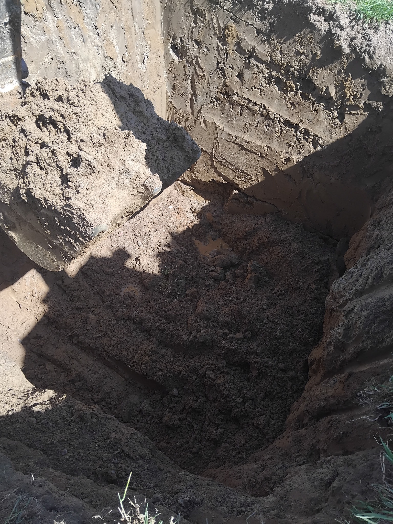 Głęboki wykop w ziemi o nierównych ścianach, oświetlony z góry, ukazujący strukturę gleby i warstwy ziemi, z fragmentami trawy na krawędzi.