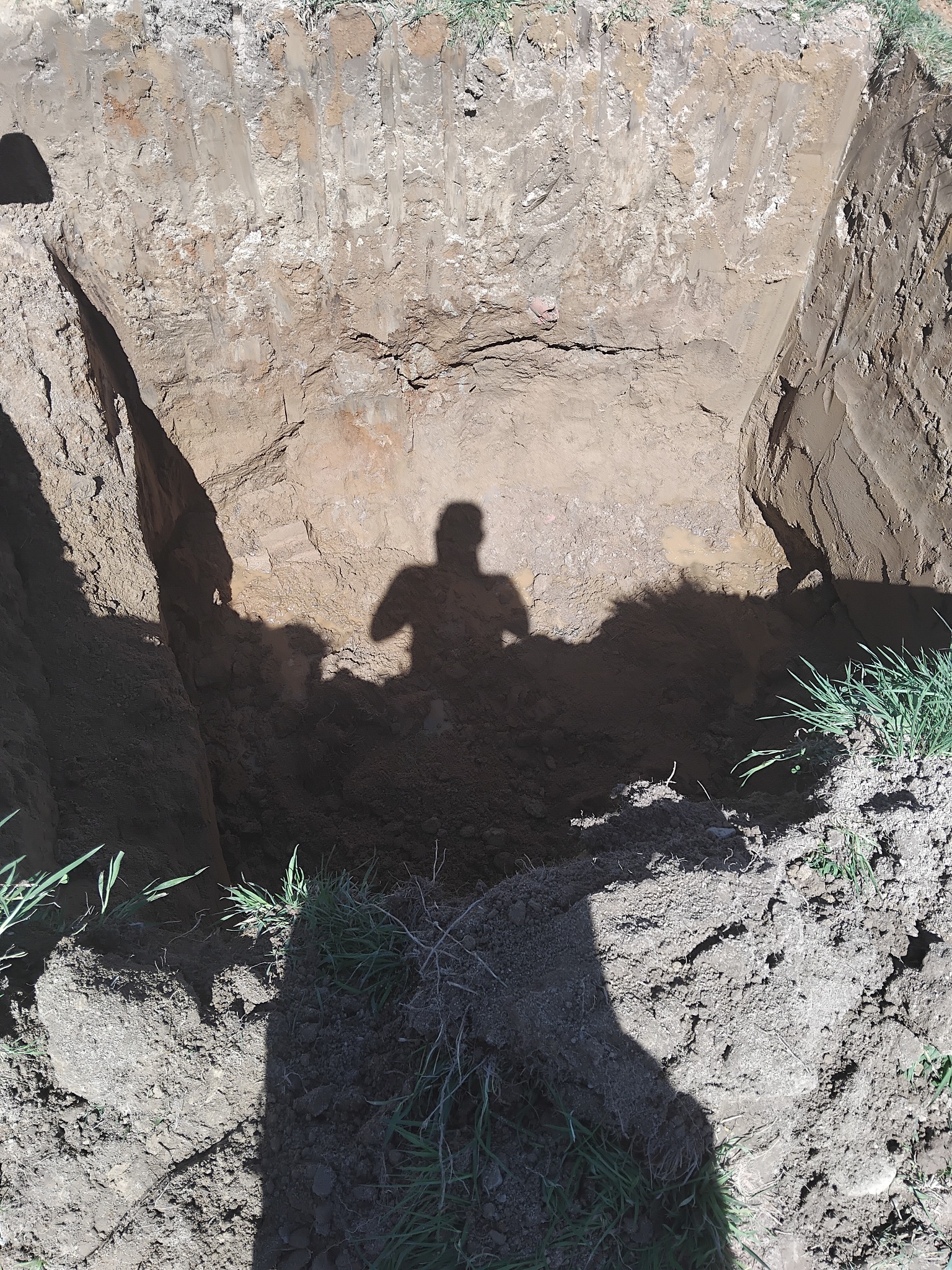 Wykop w ziemi z cieniem osoby, świeże ślady prac ziemnych. Pionowe ściany wykopu, widoczna struktura gruntu. Na górze trawa. Cień osoby na dnie wykopu.