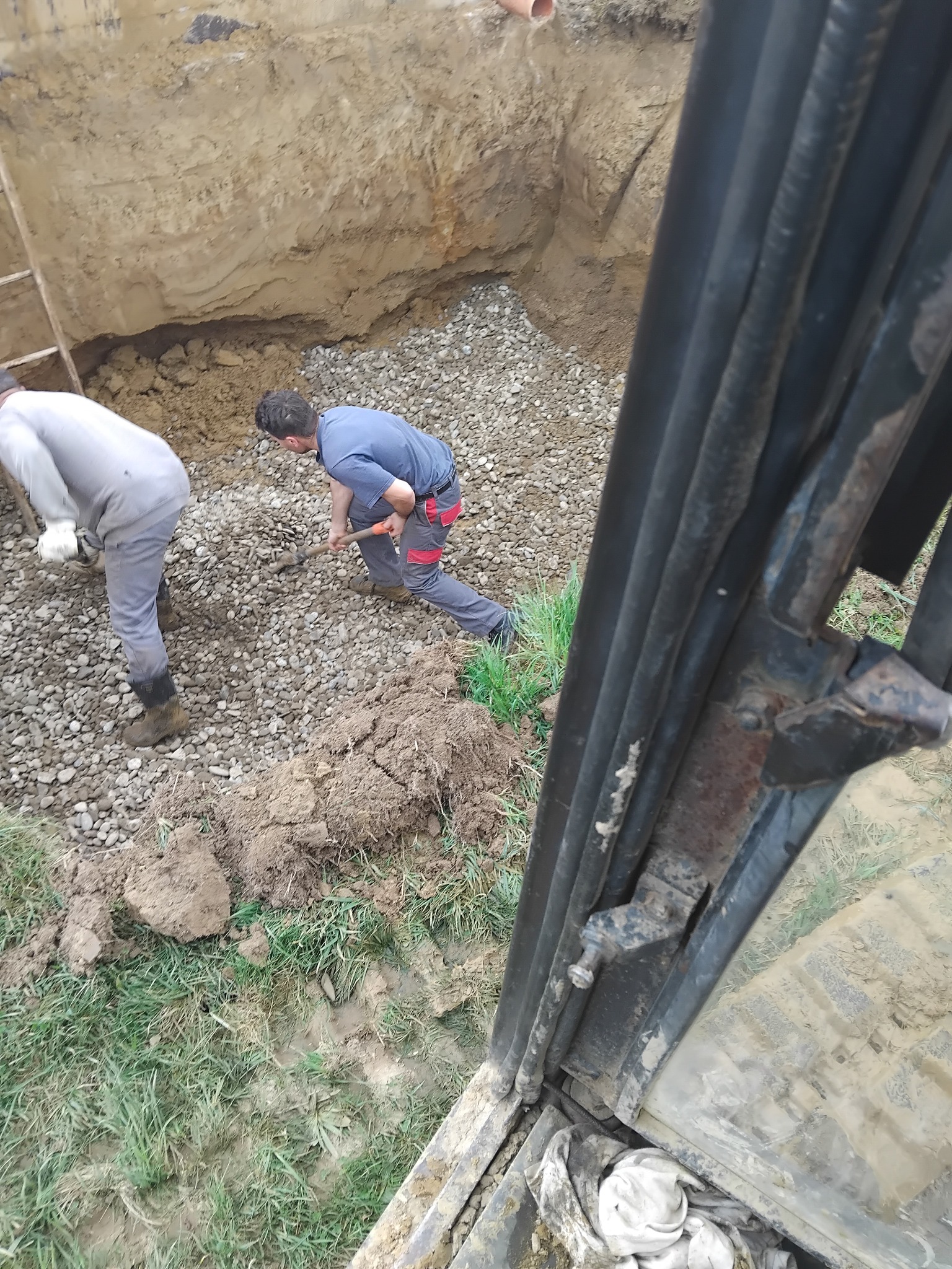 Prace ziemne w Zakliczynie: Dwóch robotników rozgarnia żwir w wykopie, widok z kabiny maszyny budowlanej. Ujęcie z góry.