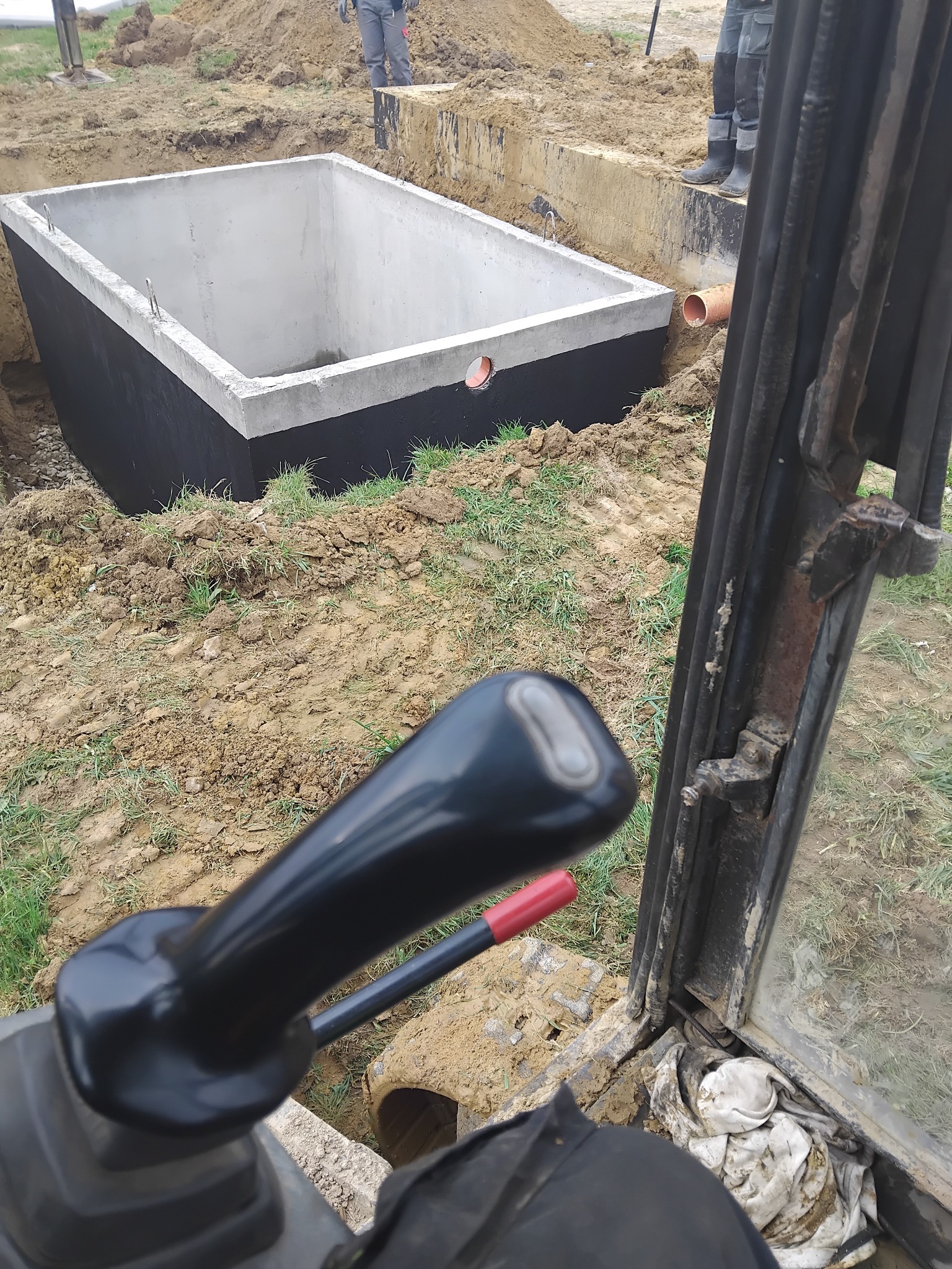 Betonowy zbiornik w wykopie, widok z kabiny koparki. Zbiornik zabezpieczony czarną powłoką, rura w ścianie. W tle ziemia i fragmenty osób.