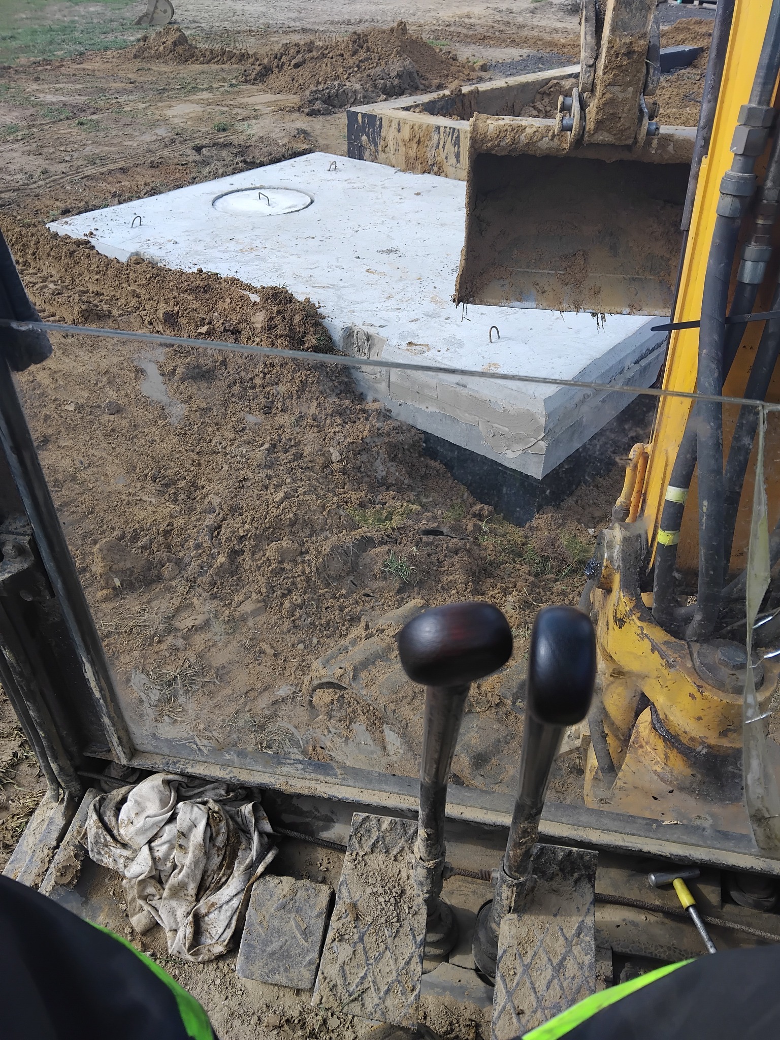 Betonowy zbiornik z włazem, częściowo zagrzebany w ziemi, widziany z kabiny koparki. Żółta łyżka koparki i dźwignie sterujące w kadrze.