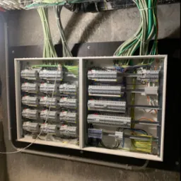 Otwarta skrzynka rozdzielcza z licznymi bezpiecznikami i przewodami w peszlach, zamontowana na surowej, szarej ścianie, z widocznymi śladami po rysunku. Nad skrzynką, pod sufitem, widoczne kable...