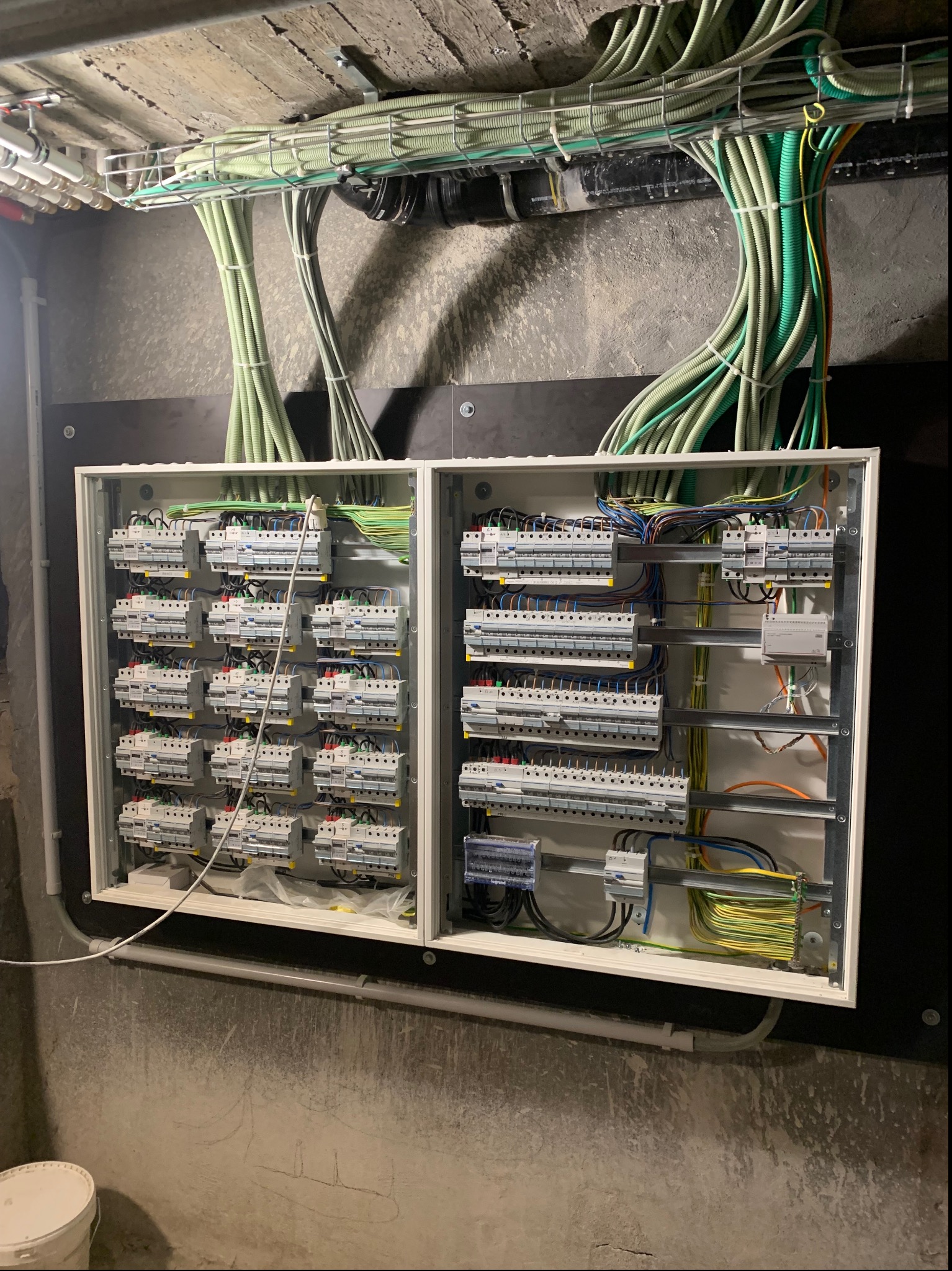 Otwarta skrzynka rozdzielcza z licznymi bezpiecznikami i przewodami w peszlach, zamontowana na surowej, szarej ścianie, z widocznymi śladami po rysunku. Nad skrzynką, pod sufitem, widoczne kable...