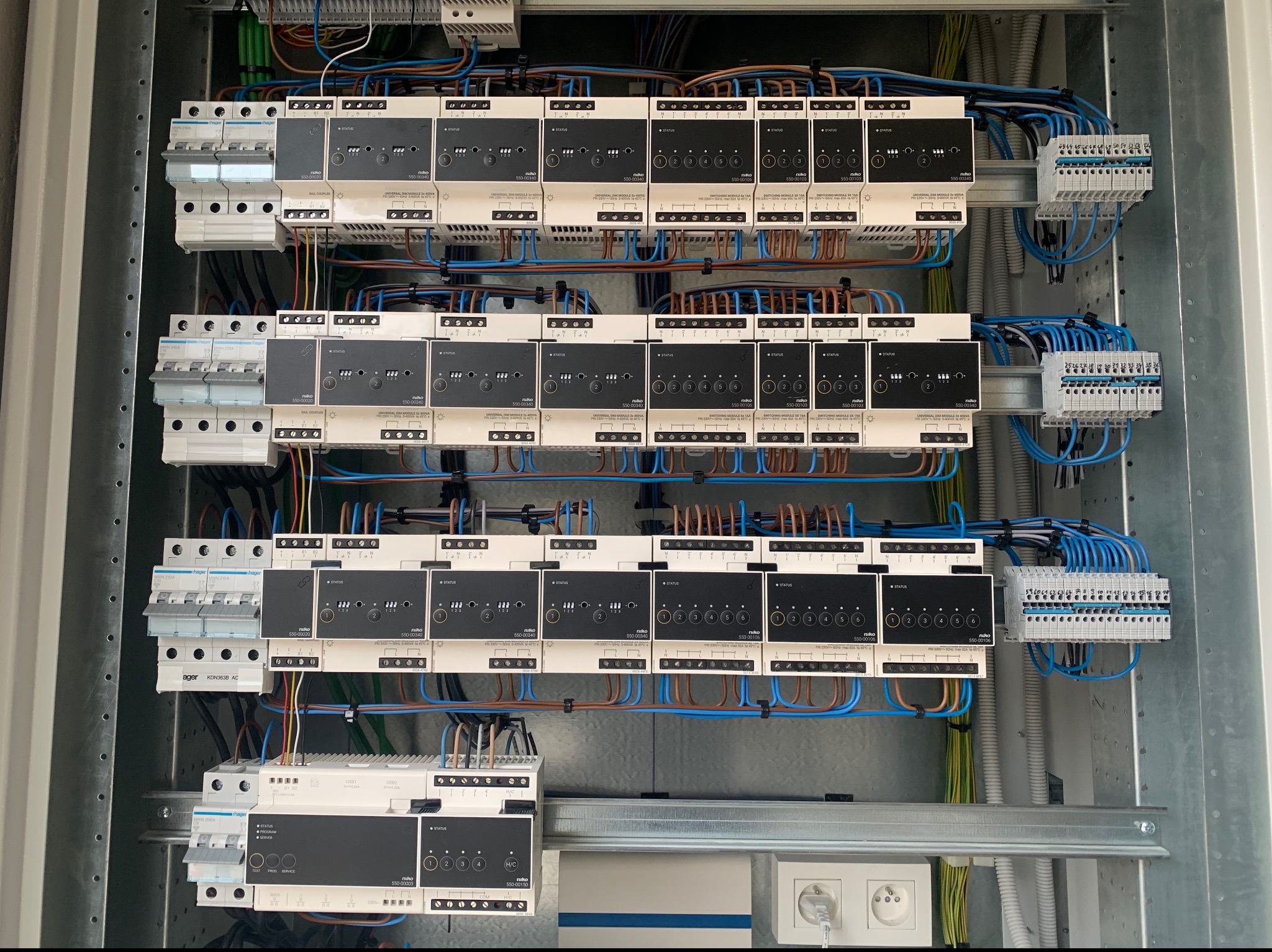 Rozdzielnica elektryczna z modułami sterującymi Niko, zabezpieczeniami Hager i okablowaniem w kolorach niebieskim i brązowym, zamontowana w metalowej obudowie.