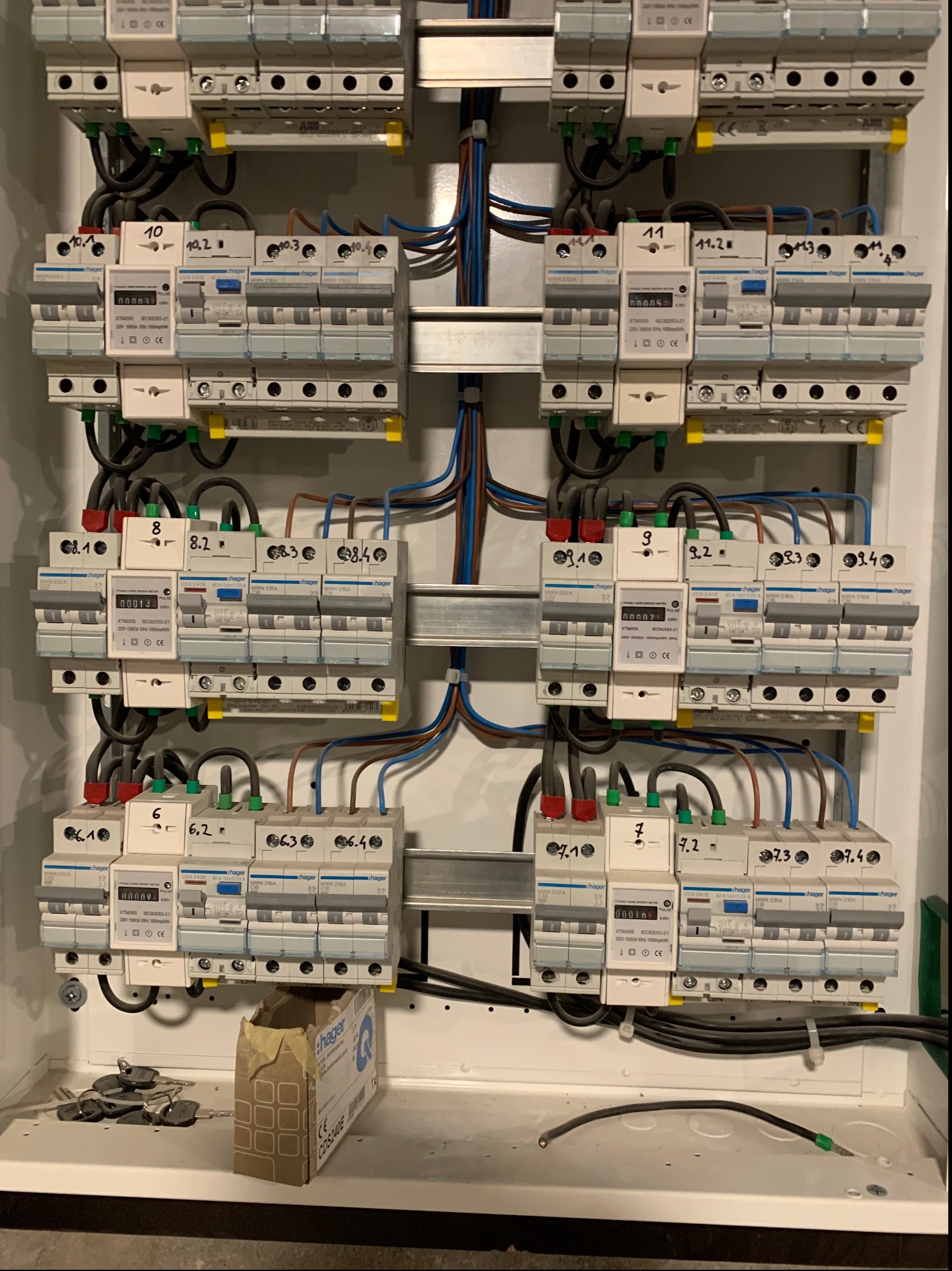 Rozdzielnica elektryczna z zamontowanymi bezpiecznikami i licznikiem energii, okablowanie uporządkowane, widoczny karton po urządzeniu Hager.