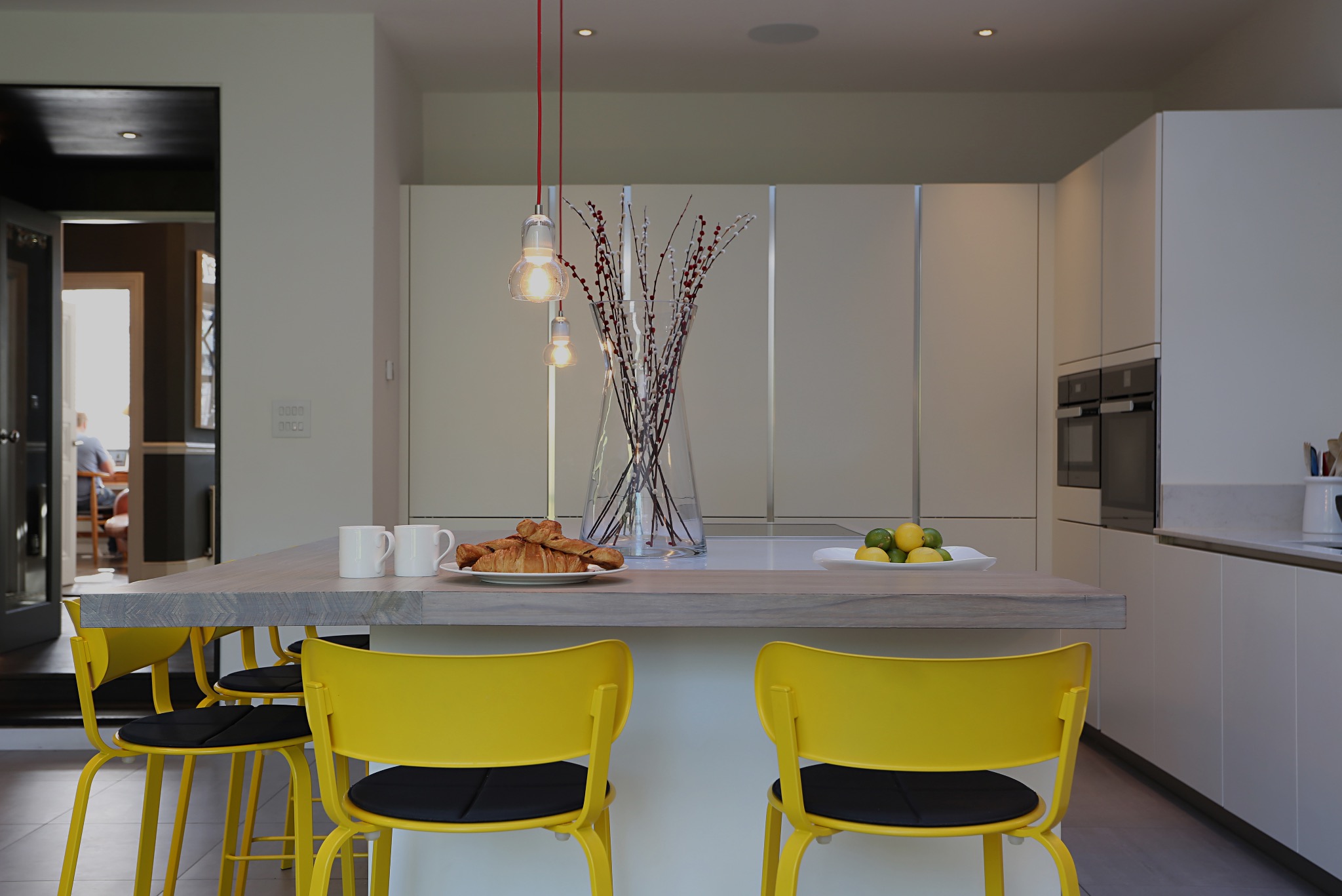 Nowoczesna kuchnia z wyspą, żółte krzesła barowe, croissanty na talerzu, minimalistyczne oświetlenie i dekoracyjne gałązki w wazonie.