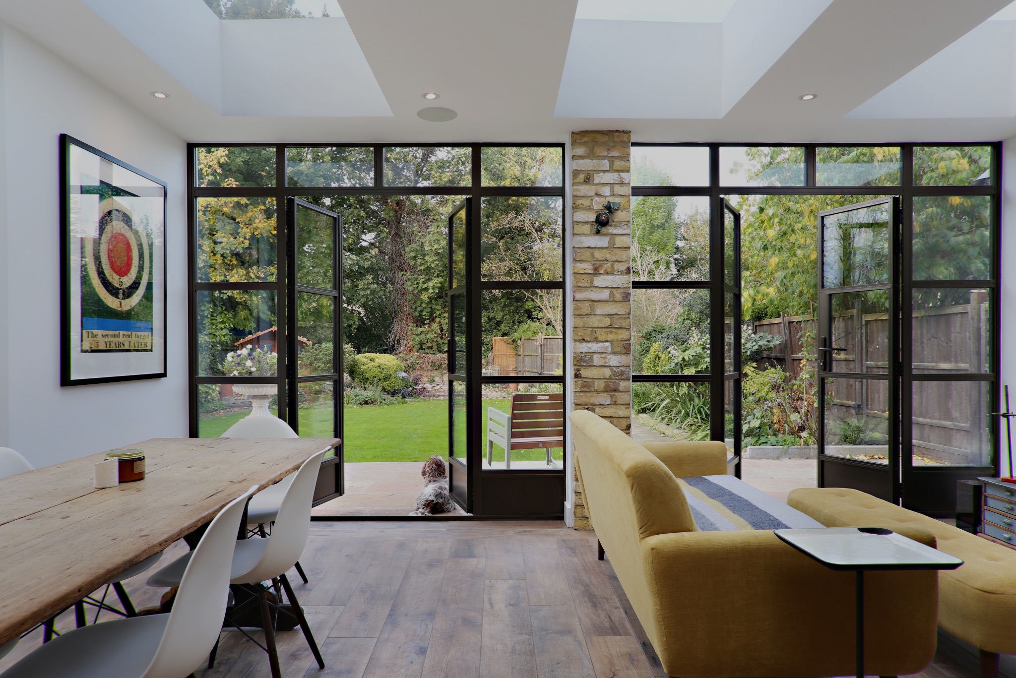 Nowoczesne wnętrze z drewnianą podłogą, żółtą sofą, stołem z białymi krzesłami i dużymi, czarnymi oknami wychodzącymi na ogród, przez które widać psa