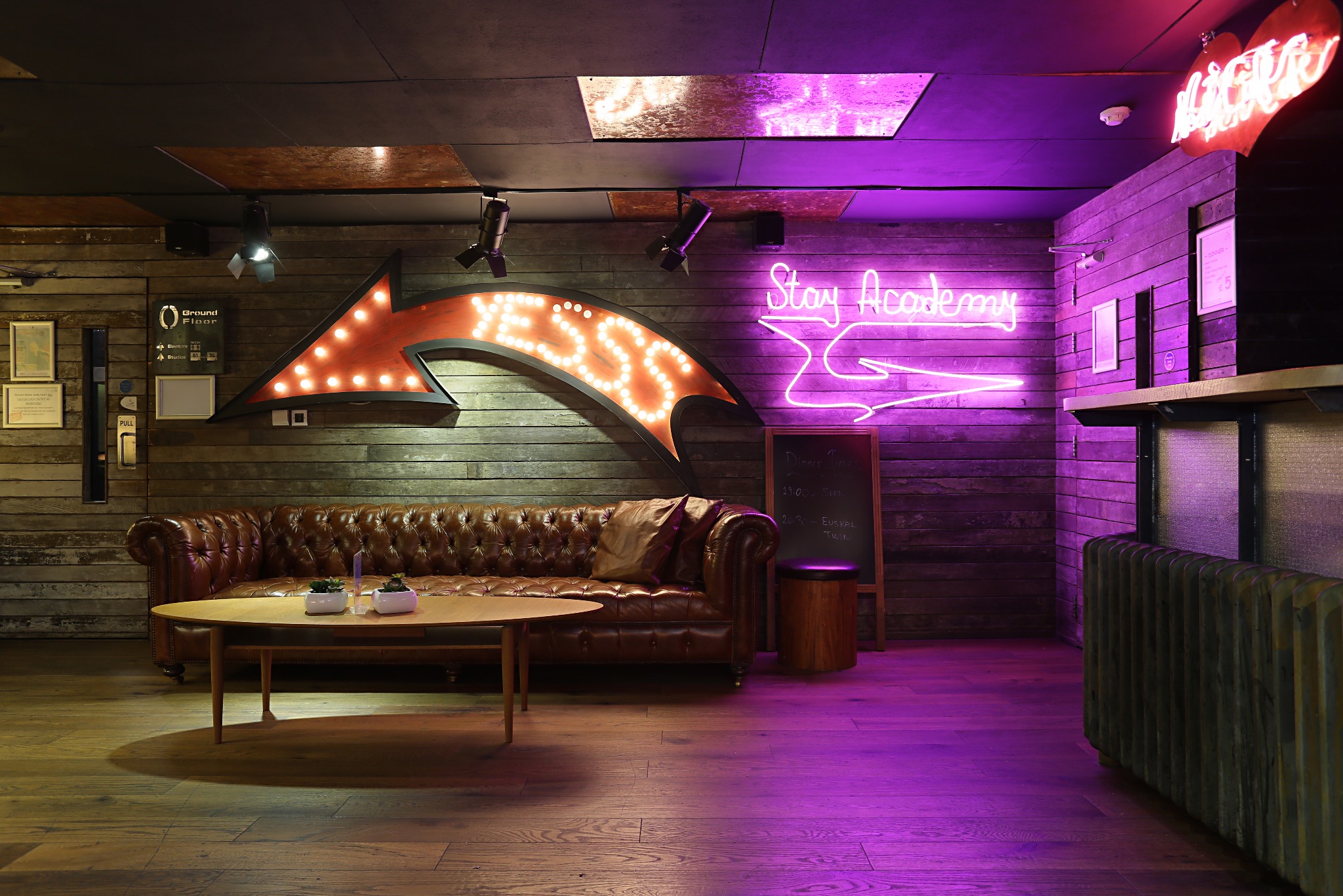 Rustykalne wnętrze z brązową skórzaną sofą Chesterfield, okrągłym drewnianym stolikiem kawowym, neonowym napisem 'Stay Academy' i strzałką z żarówkami na drewnianej ścianie, oświetlone fioletowym...